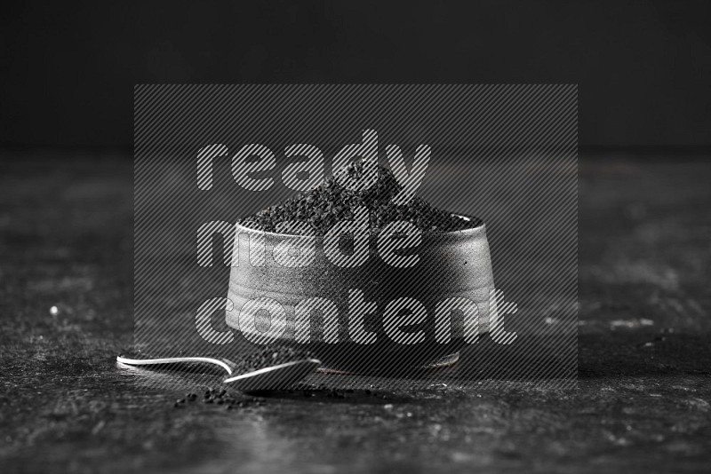 A black pottery bowl full of black seeds with a metal spoon full of the seeds on a textured black flooring