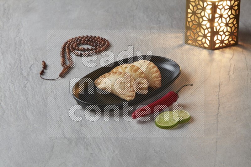Three fried sambosas in an oval shaped black plate and a red sauce in a black round ramekin with a brown misbaha and a golden lantern on the side on a gray background