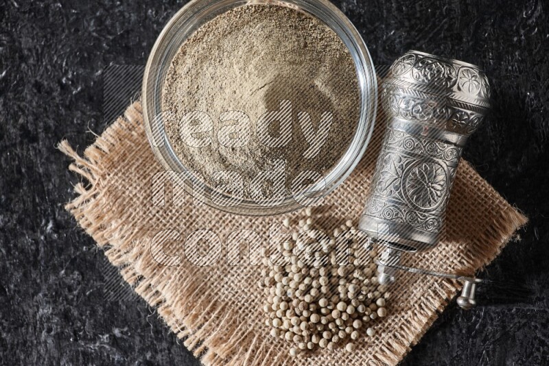 A glass bowl full of white pepper powder with white pepper beads on a burlap piece of fabric and a metal grinder on textured black flooring
