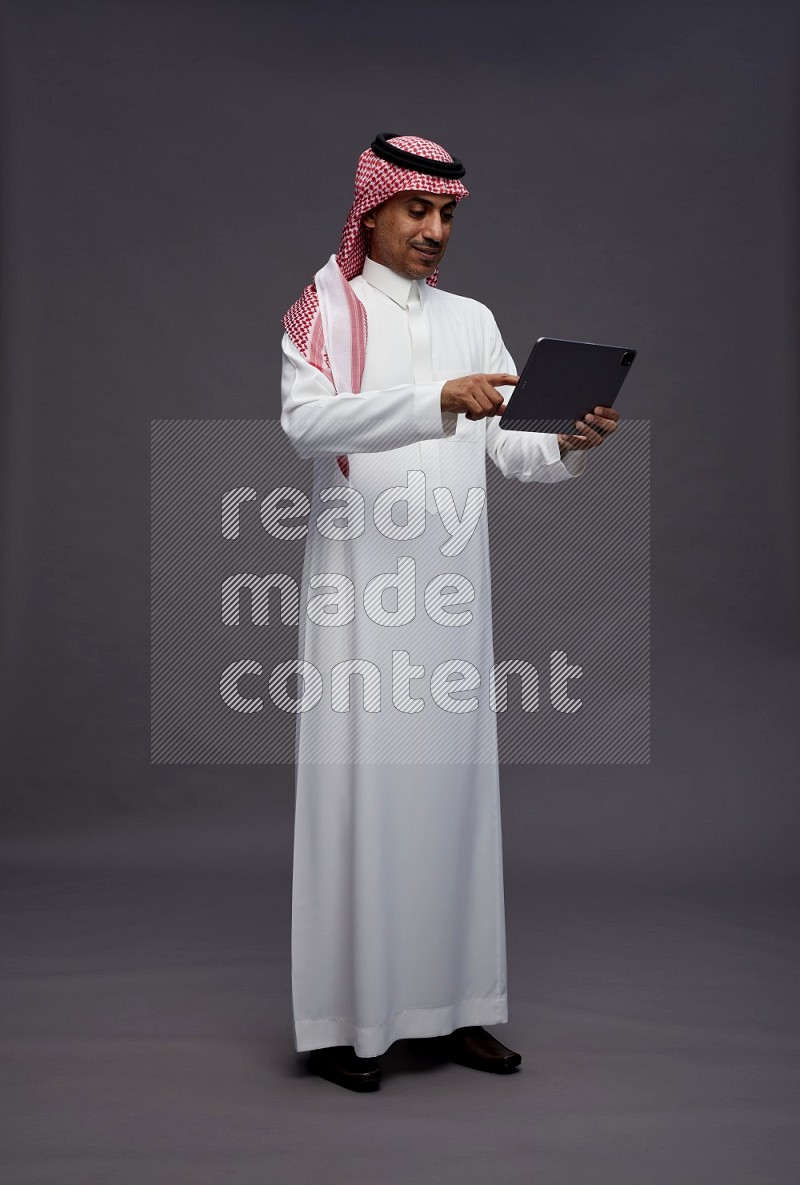 Saudi man wearing thob and shomag standing working on tablet on gray background