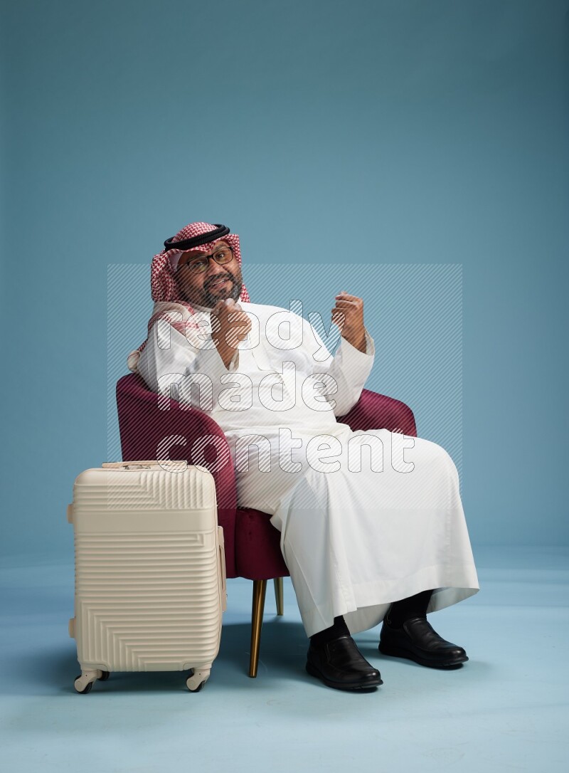 Saudi Man with shimag sitting on chair with a travel luggage on blue background