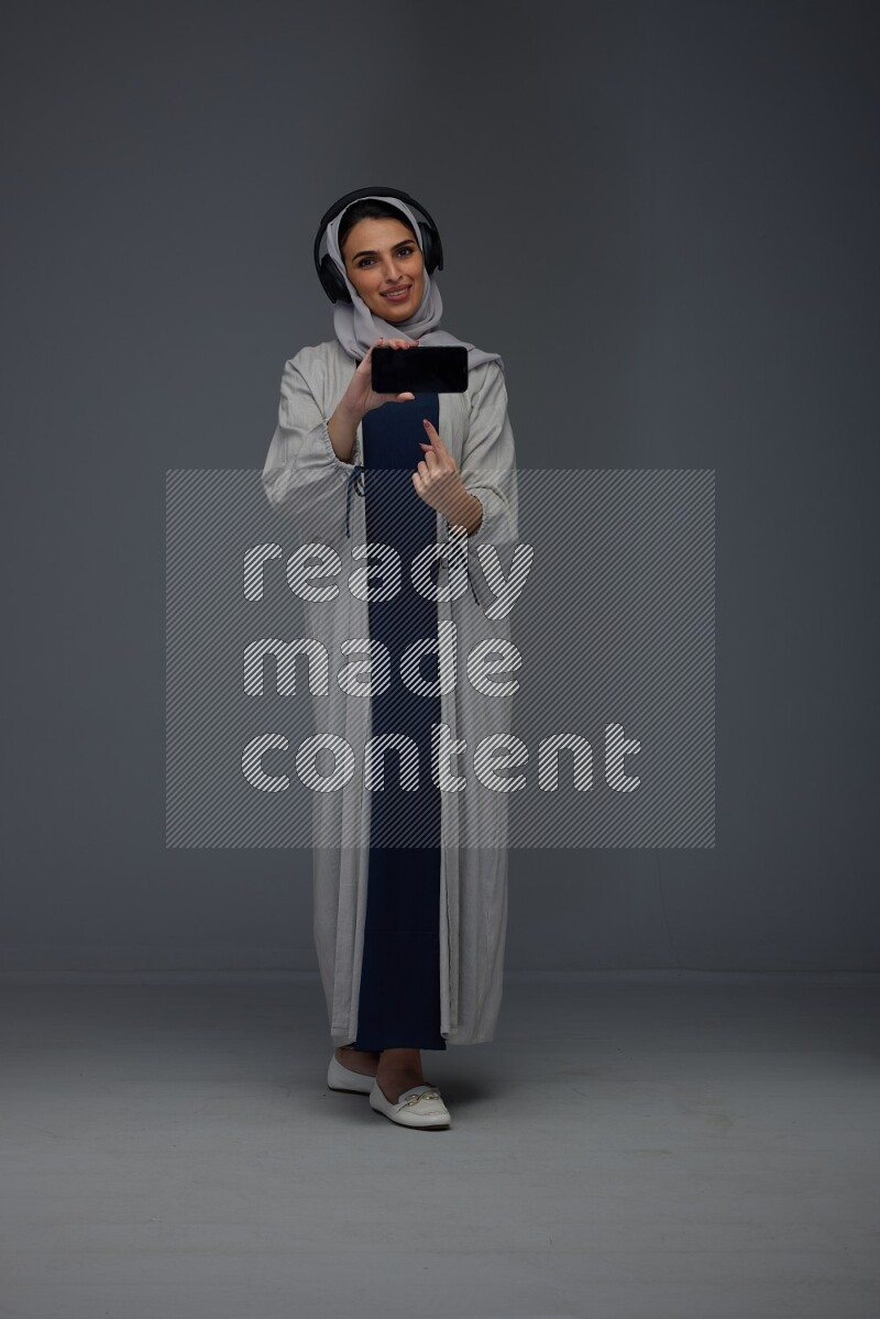 A Saudi woman wearing a light gray Abaya and head scarf setting on a dark grey chair and watching on the tablet eye level on a grey background