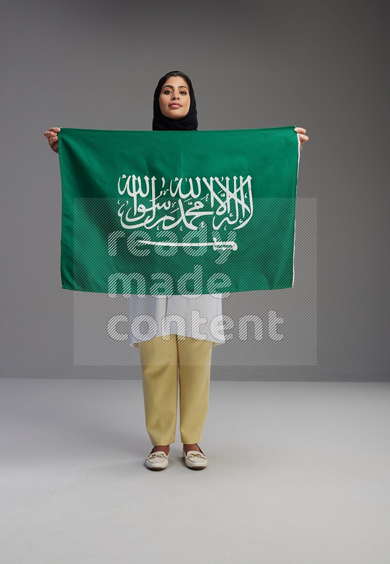 Saudi woman wearing lab coat with stethoscope standing holding Saudi flag on Gray background
