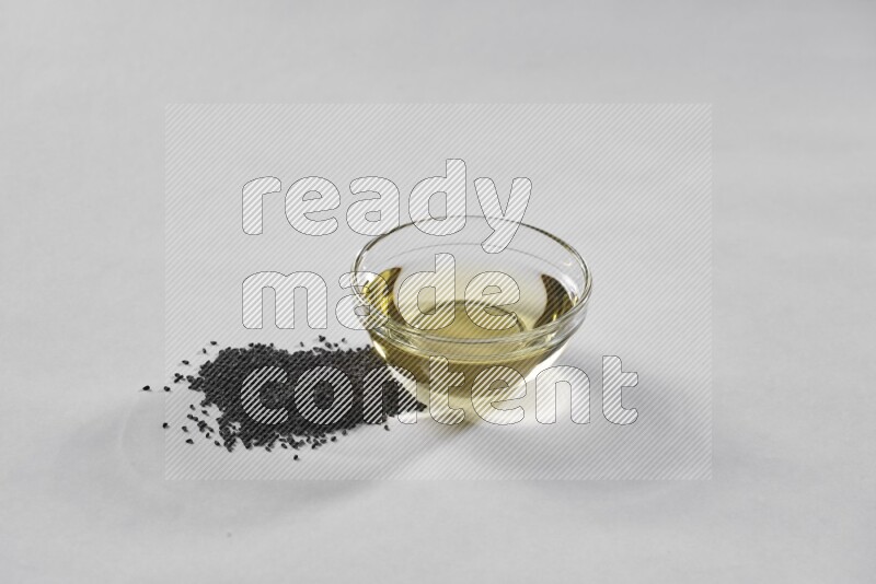 A glass bowl full of black seeds oil with black seeds beside it on a white flooring