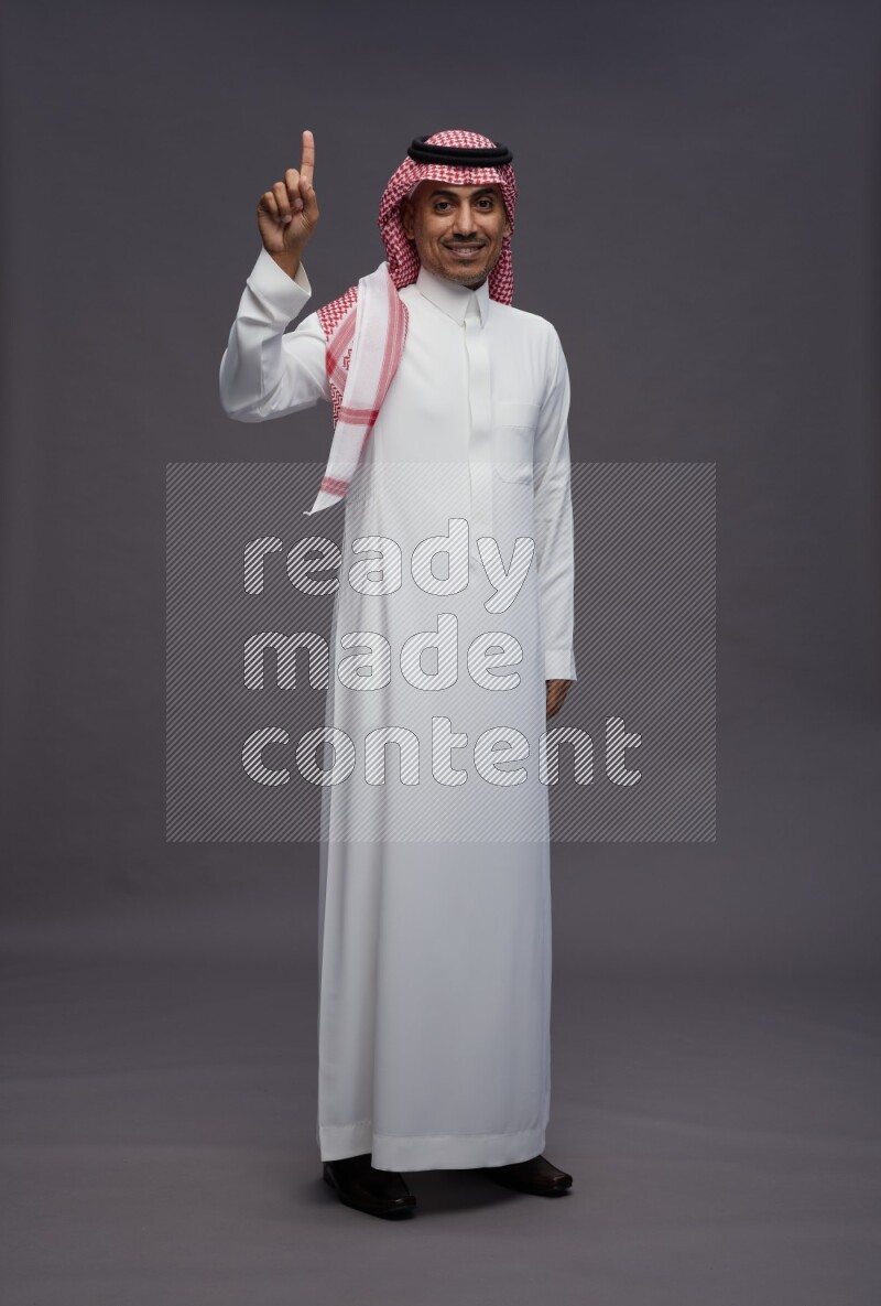 Saudi man wearing thob and shomag standing interacting with the camera on gray background