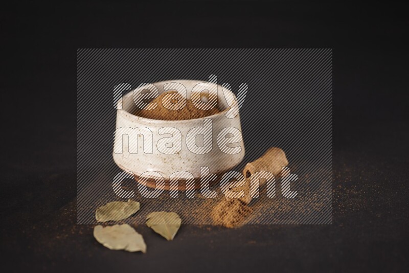 Cinnamon powder in a white pottery bowl and cinnamon sticks and laurel leaves on black background