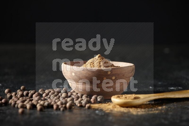 A wooden bowl and spoon full of allspice powder and whole balls spreaded on a black flooring