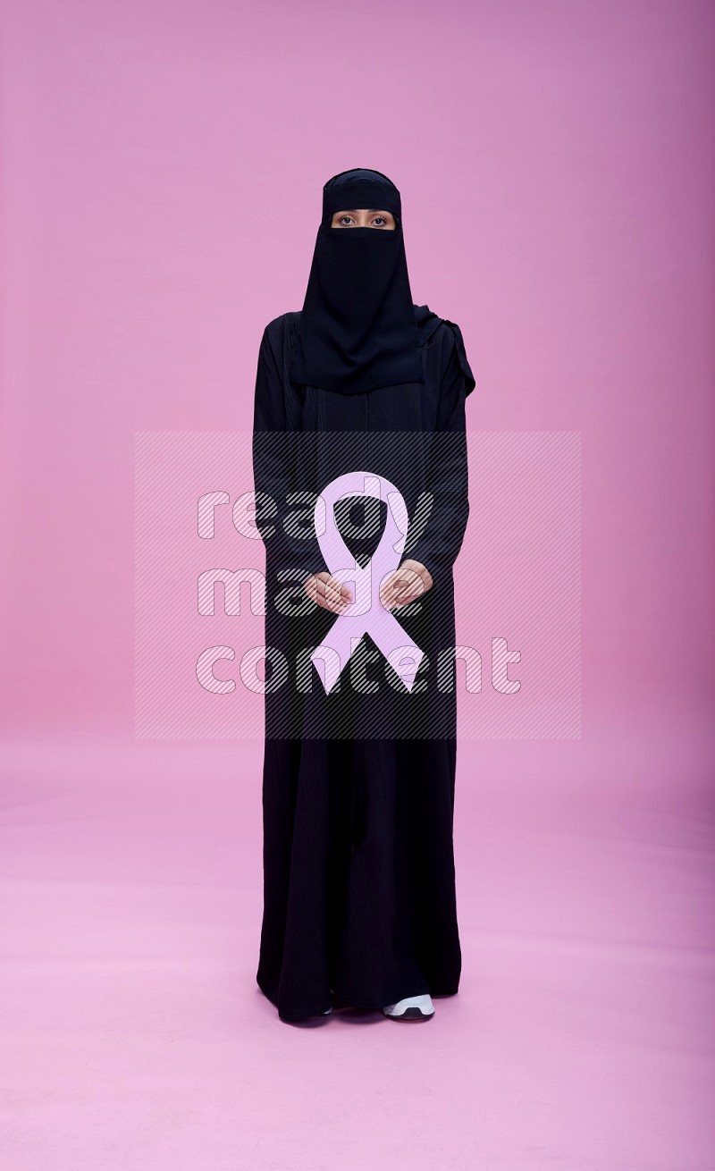 Saudi woman wearing abaya and niqab standing holding awareness ribbon on plain pink background
