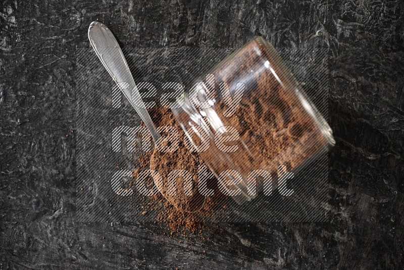 A flipped glass jar and metal spoon full of cloves powder with cloves spread on a textured black flooring