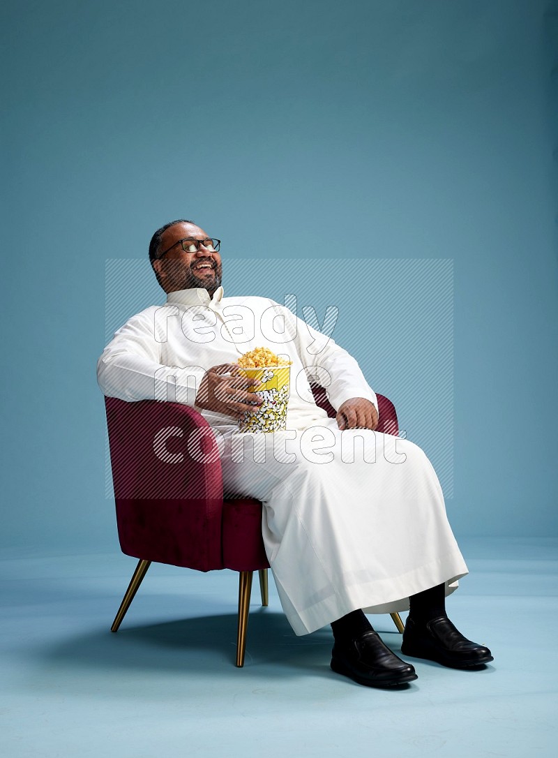 Saudi Man without shimag sitting on chair eating popcorn on blue background