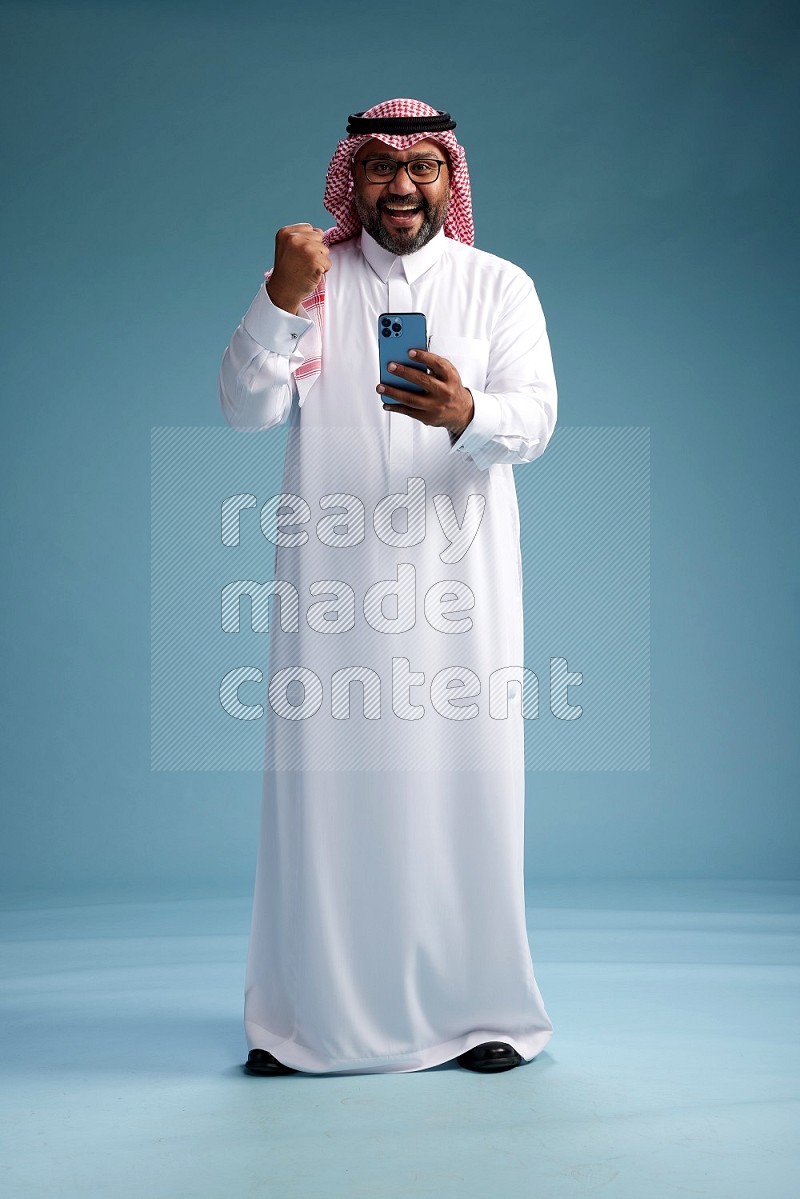 Saudi Man with shimag Standing texting on phone on blue background