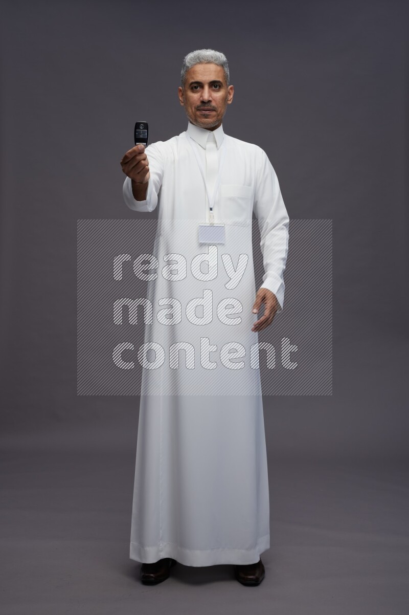 Saudi man wearing thob with neck strap employee badge standing holding key car on gray background