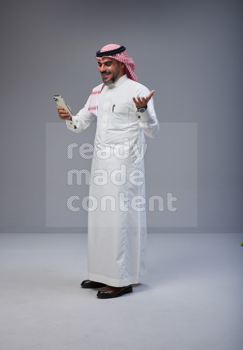 Saudi man Wearing Thob and red Shomag standing texting on phone on Gray background