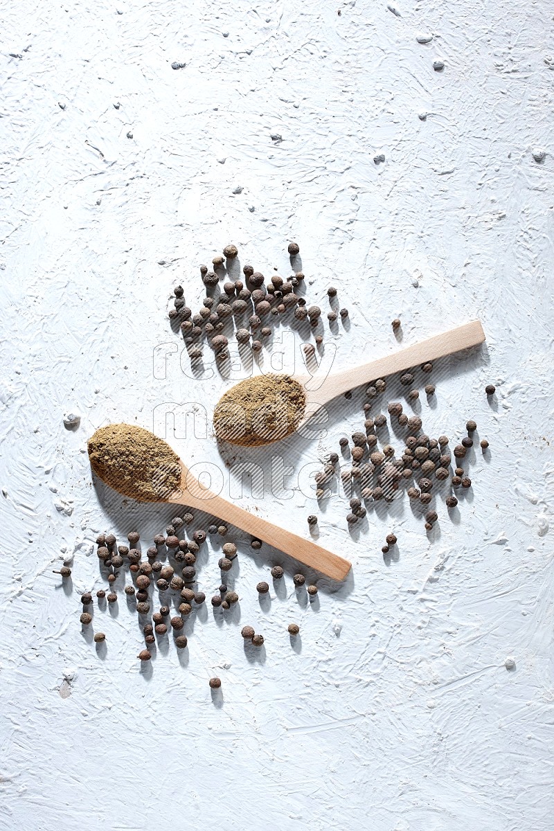 Wooden spoons full of allspice powder and allspice whole balls spreaded on a textured white flooring