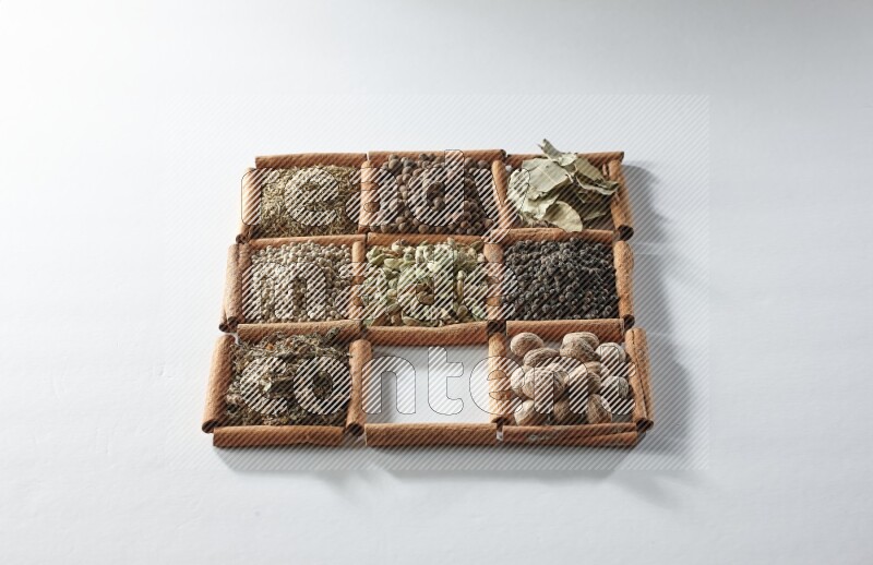 9 مربعات من أعواد القرفة ممتلئة بالهيل في المنتصف محاطة بجوزة الطيب والملح والريحان المجفف والفلفل الأبيض والكمون وبهار الفلفل الحلو وأوراق الغار والفلفل الأسود على أرضية بيضاء