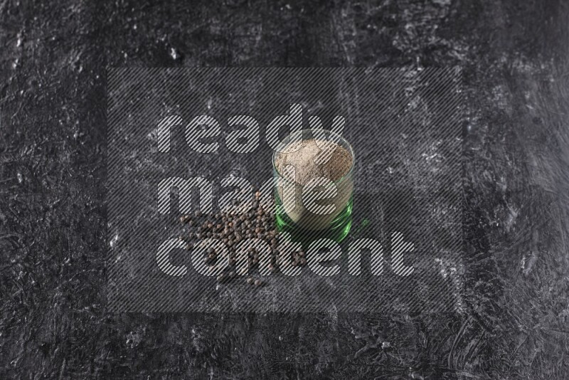 A glass cup full of black pepper powder with beads beside it on a textured black flooring