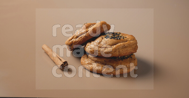 Three chocolate chip cookies beside cinimon sticks on a brown background