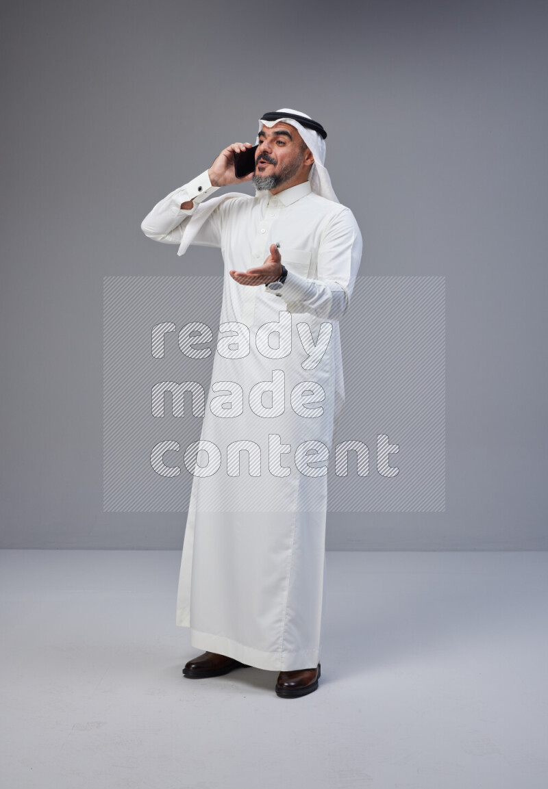 Saudi man Wearing Thob and white Shomag standing talking on phone on Gray background