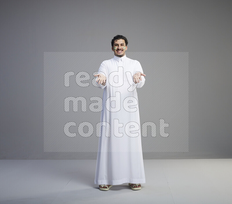 A Saudi man standing wearing thob with face painting interacting with the camera on gray background