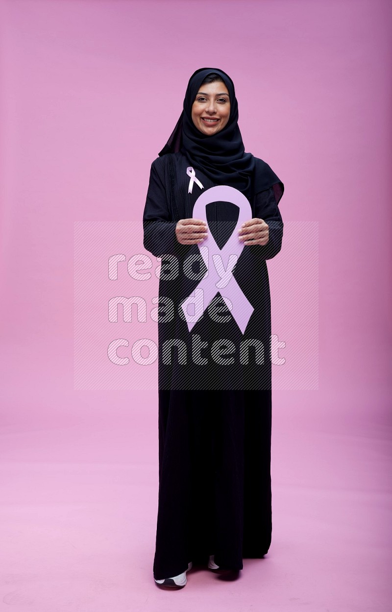 Saudi woman wearing pink ribbon on abaya standing holding awareness ribbon on plain pink background