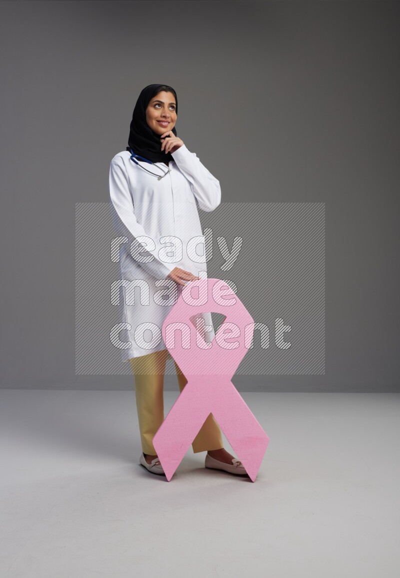 Saudi woman wearing lab coat with stethoscope standing holding awareness ribbon symbol standing on Gray background