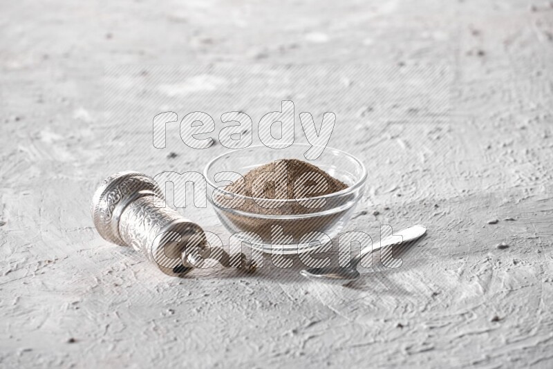 A glass bowl full of black pepper powder and a metal spoon and a turkish metal grinder on textured white flooring