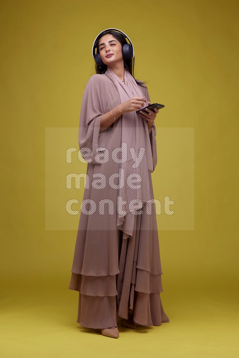 A Saudi woman Listing to Music on a Yellow Background wearing Brown Abaya