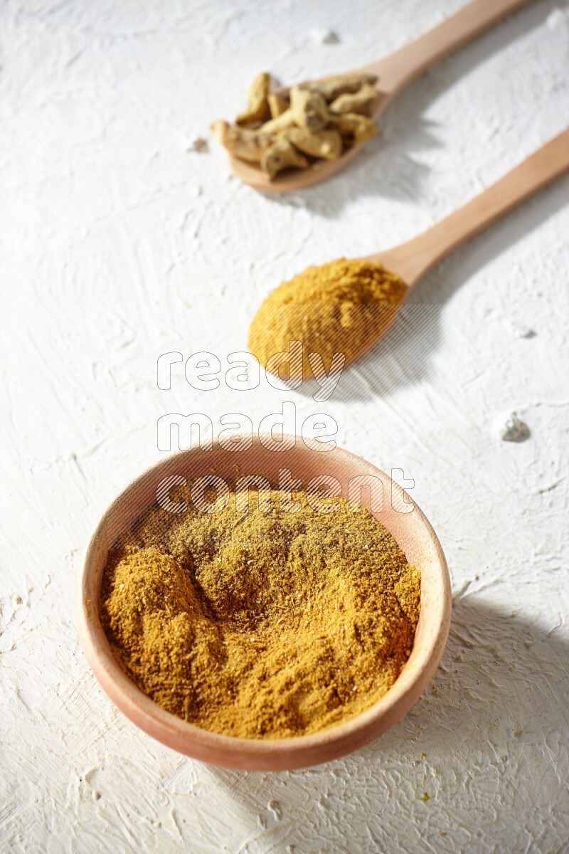 A wooden bowl full of turmeric powder and 2 wooden spoons full of dried turmeric whole finger and turmeric powder on textured white flooring
