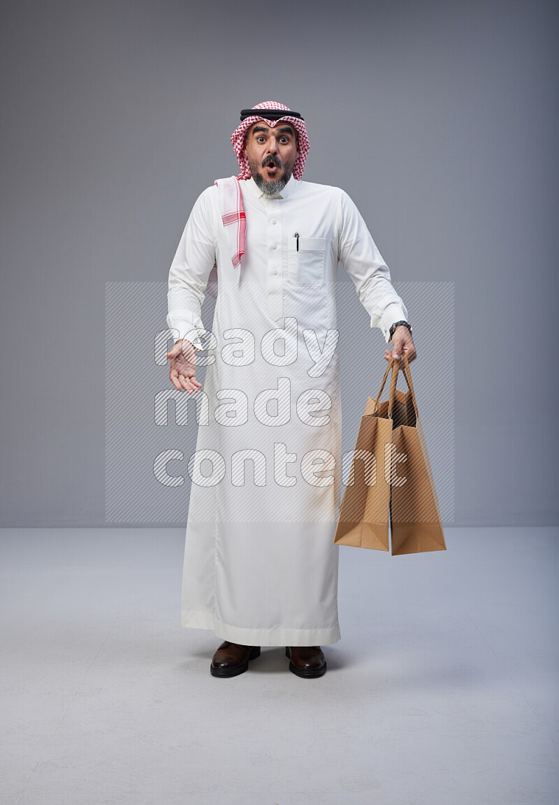 Saudi man Wearing Thob and red Shomag standing holding shopping bag on Gray background