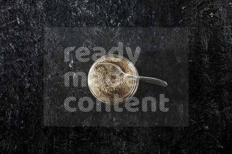 A glass jar and metal spoon full of cardamom powder on textured black flooring