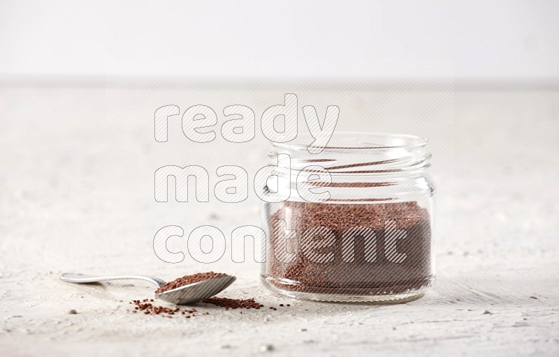A glass jar and a metal spoon full of garden cress on a textured white flooring in different angles