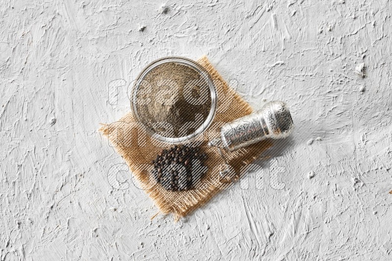 A glass bowl full of black pepper powder, black pepper beads and a turkish metal grinder on burlap fabric on textured white flooring