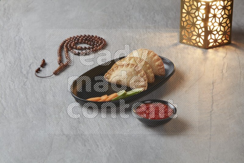Three fried sambosas in an oval shaped black plate and a red sauce in a black round ramekin with a brown misbaha and a golden lantern on the side on a gray background