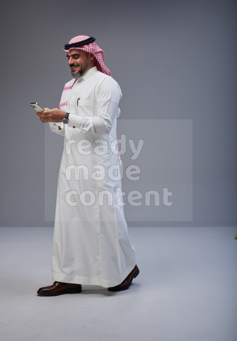Saudi man Wearing Thob and red Shomag standing texting on phone on Gray background