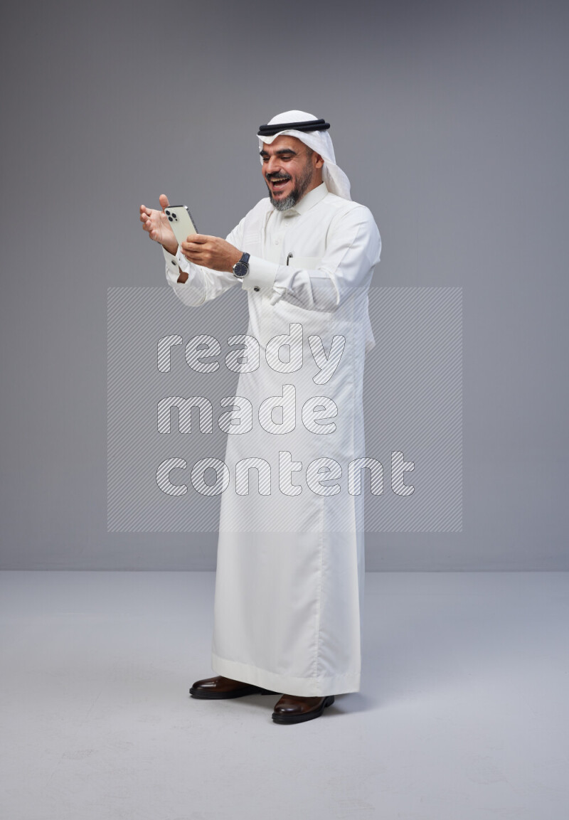 Saudi man Wearing Thob and white Shomag standing texting on phone on Gray background