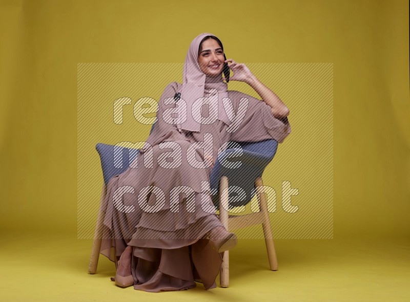 A woman having a call on a blue Chair on a Yellow Background wearing Brown Abaya with Hijab