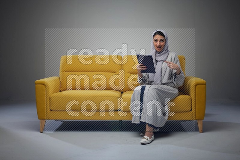 A Saudi woman wearing a light gray Abaya and head scarf sitting on a yellow sofa and holding her tablet while pointing to it eye level on a grey background