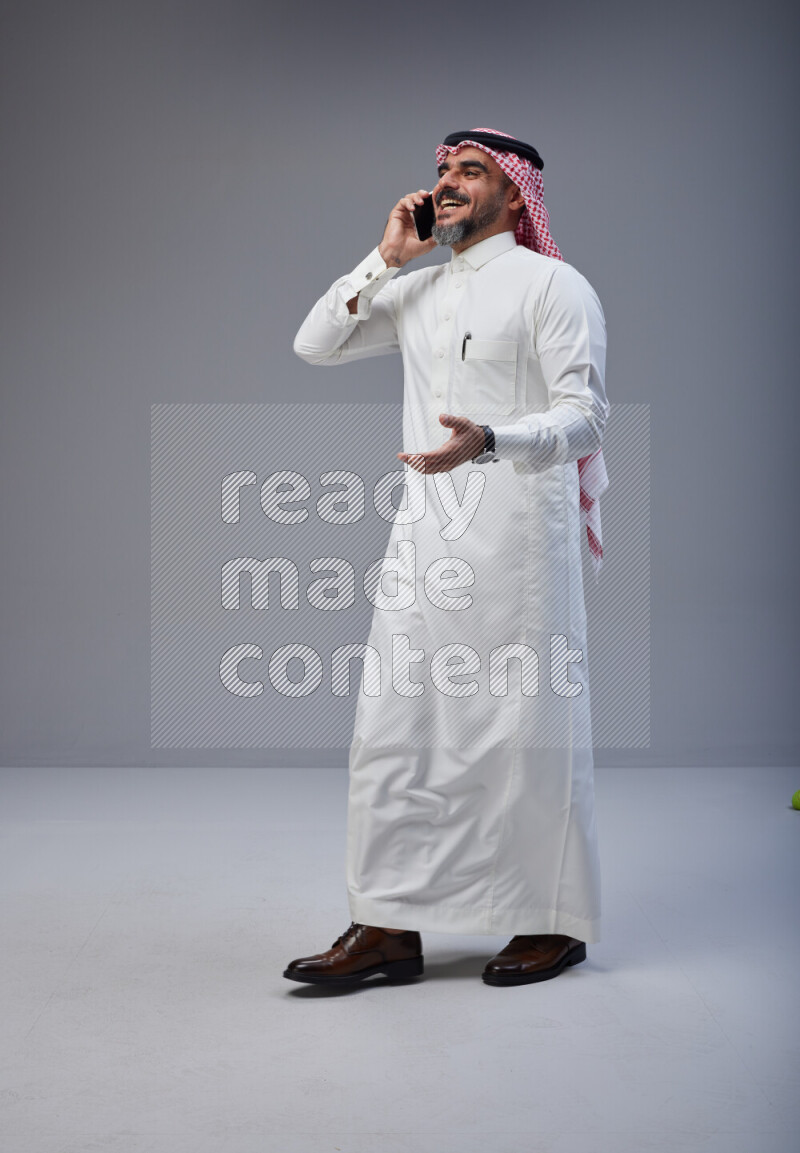 Saudi man Wearing Thob and red Shomag standing talking on phone on Gray background