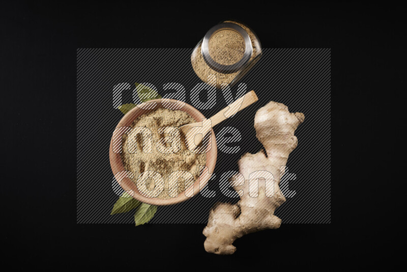 A wooden bowl full of ground ginger powder with a glass jar beside it and fresh ginger on black background