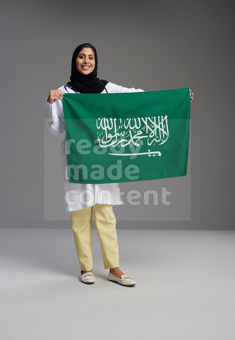 Saudi woman wearing lab coat with stethoscope standing holding Saudi flag on Gray background