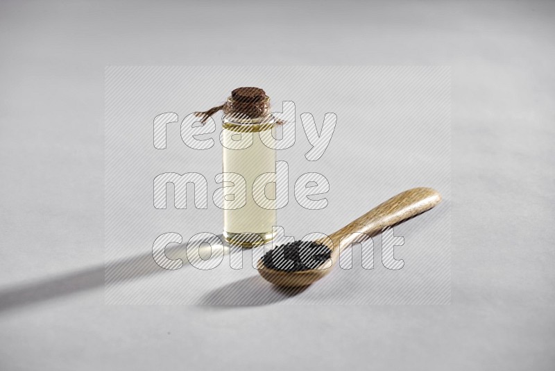 A wooden spoon full of black seeds with a glass bottle of black seeds oil on a white flooring