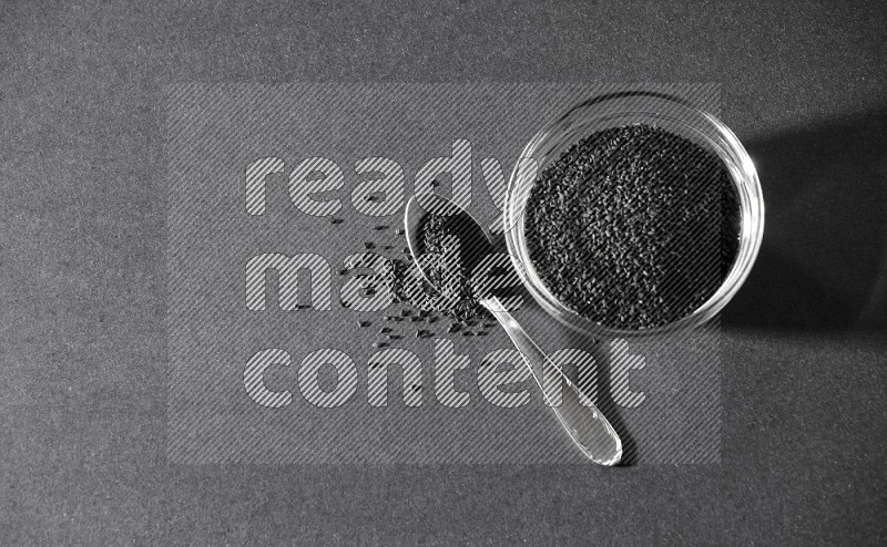 A glass bowl full of black seeds with a metal spoon full of the seeds on a black flooring