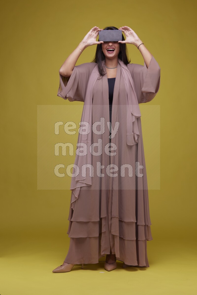 A woman Showing SmartPhone Screen on a Yellow Background wearing Brown Abaya