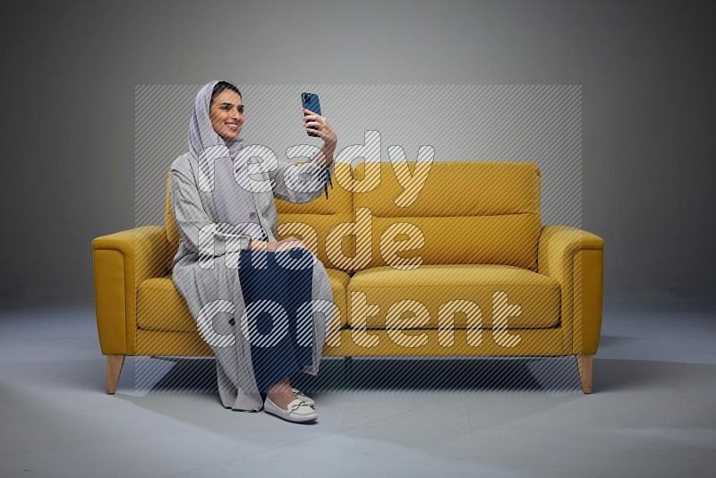 A Saudi woman wearing a light gray Abaya and head scarf sitting on a yellow sofa and making a video call eye level on a grey background