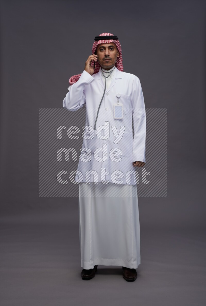 Saudi man wearing thob with lab coat and shomag with pocket employee badge standing talking on phone on gray background