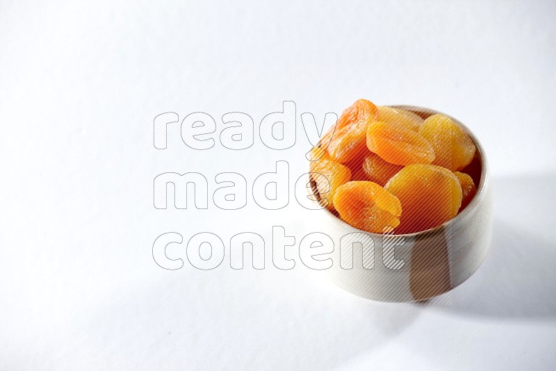 A beige ceramic bowl full of dried apricots on a white background in different angles