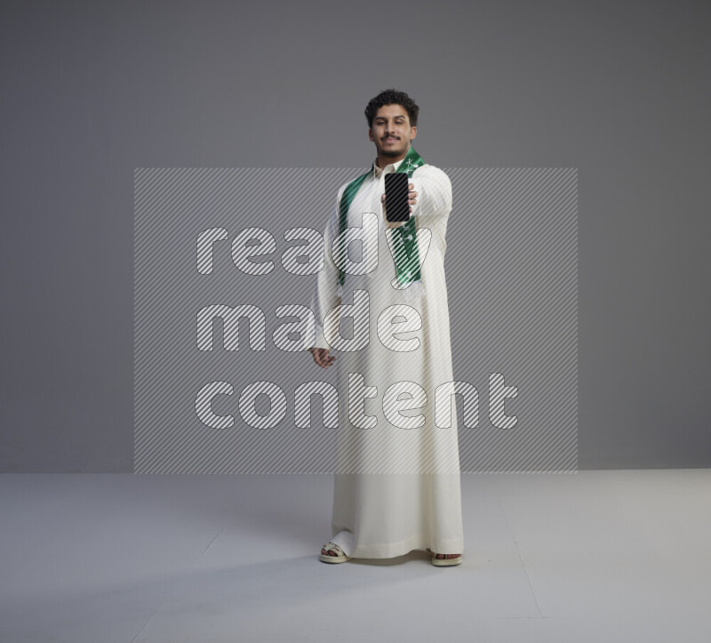 A Saudi man standing wearing thob and Saudi flag scarf showing phone to camera on gray background