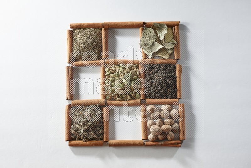 9 مربعات من أعواد القرفة ممتلئة بالهيل في المنتصف محاطة بجوزة الطيب والملح والريحان المجفف والفلفل الأبيض والكمون وبهار الفلفل الحلو وأوراق الغار والفلفل الأسود على أرضية بيضاء