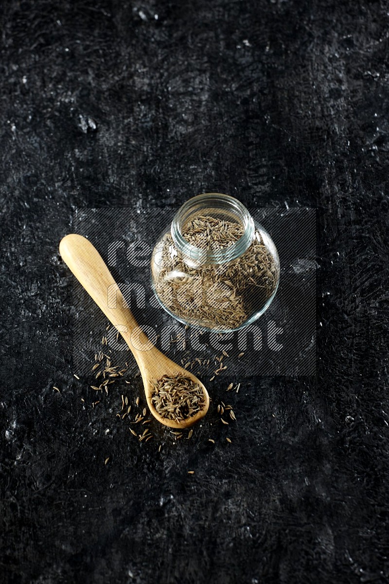 وعاء زجاجي للتوابل وملعقة خشبية ممتلئين بحبوب الكمون على خلفية سوداء
