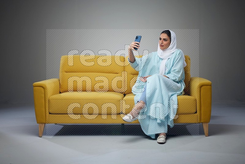 A Saudi woman wearing a light blue Abaya and white head scarf sitting on a yellow sofa and taking selfies with her phone eye level on a grey background